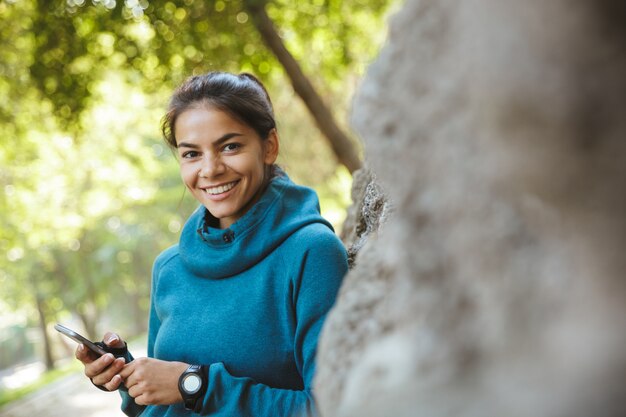 Close-up de uma mulher jovem e atraente fitness vestindo roupas esportivas, fazendo exercícios ao ar livre, usando telefone celular