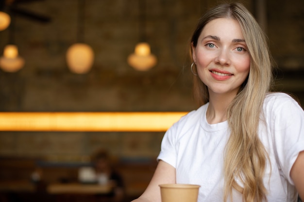 Close-up de uma mulher loira sorridente em uma camiseta branca que está sentado em um café com um copo de papel. lugar para o seu design