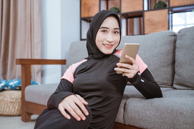 Close-up de uma mulher vestindo uma roupa esportiva hijab sorrindo ao usar um telefone celular enquanto está sentada relaxadamente em um tapete de ioga, enquanto se inclina no sofá