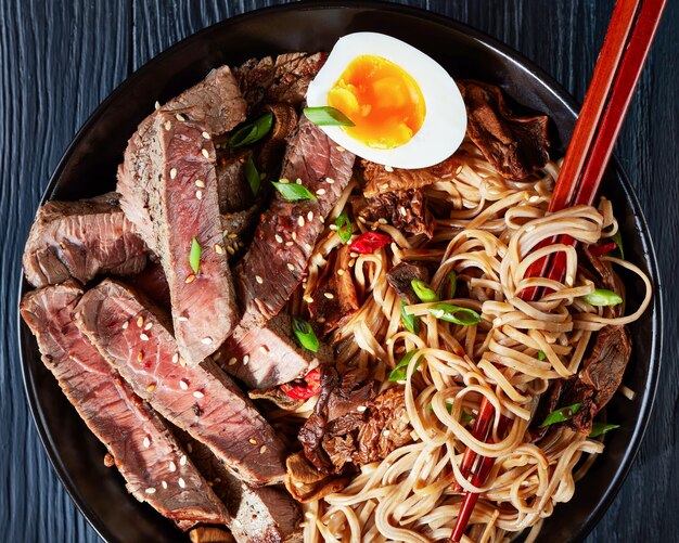 Close-up de uma tigela preta de macarrão Soba com rosbife fatiado, cogumelos shiitake e metade de ovo cozido