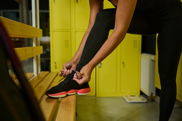 Close-up de una deportista irreconocible atarse los cordones de los zapatos poniendo su pie sobre un banco de madera en el vestuario del gimnasio