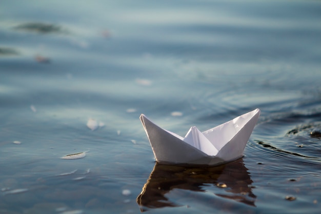 Close-up do barco de papel origami branco pequeno simples que flutua quietamente no rio ou na água do mar clara azul sob o céu brilhante do verão. Conceito de liberdade, sonhos e fantasias, fundo copyspace.