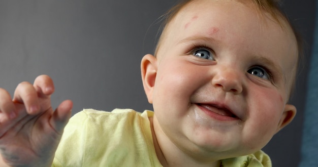 Close up do bebê encantador com olhos azuis olhando para longe com sorriso animado no pano de fundo cinza