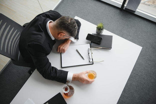 Close-up do empresário estressado segurando um copo de uísque, ele dormindo e documento de negócios de gráficos de dados na mesa do escritório, vício em álcool e conceito de empresário bêbado