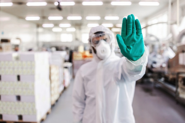 Close-up do homem em uniforme de proteção com luvas de borracha em pé na fábrica de alimentos e mostrando o sinal de stop com a mão. conceito de surto de vírus corona.