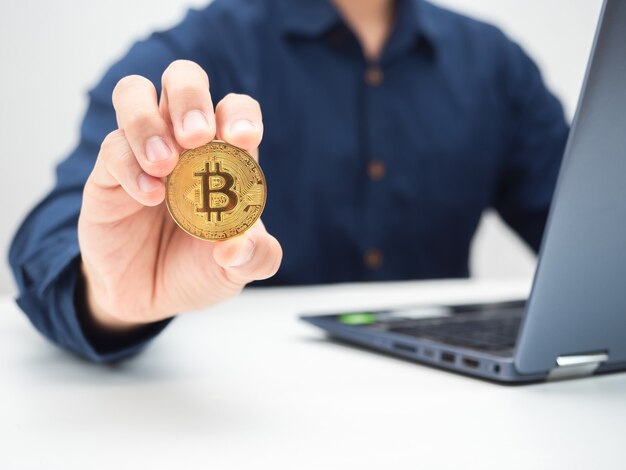 Close-up do homem mostrando bitcoin dourado na mesa com foto do laptop