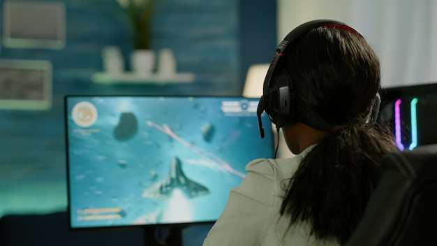 Close-up do jogador competitivo de mulher negra segurando o joystick jogando videogame de atirador espacial no poderoso computador tarde da noite. jogador profissional usando controlador sem fio para campeonato de jogos online