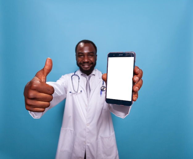 Close-up do médico sorridente desistindo de polegares enquanto segura o smartphone com tela branca sobre fundo azul. médico profissional de saúde com estetoscópio aprovando maquete em dispositivo touchscreen