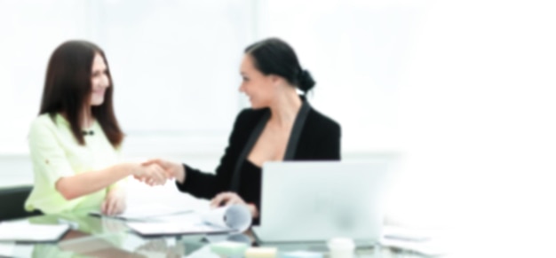 Close up. Duas mulheres de negócios apertando as mãos uma da outra, sentadas à mesa no escritório. Plano de fundo para texto com pessoas desfocadas