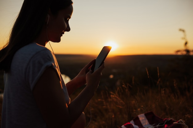 Close-up e vista lateral da mulher usando o telefone durante o pôr do sol lindo.