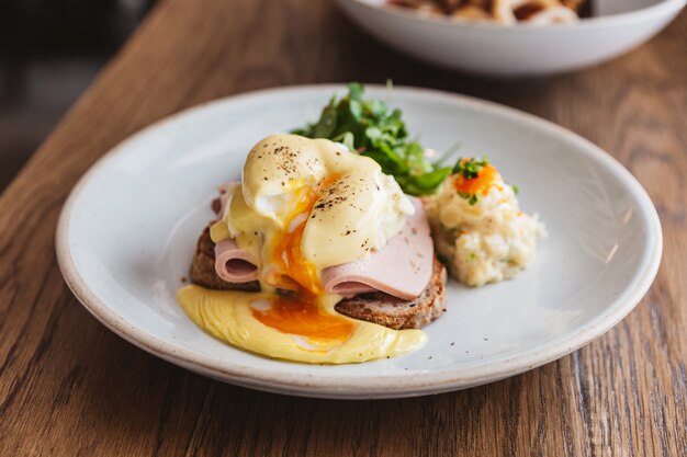 Close-up Eggs Benedict mit Schinken, der filmisches Eigelb und Käse, Toast und Kartoffelpüree schneidet und zeigt. Serviert mit Salat auf einem weißen Teller auf Holztisch.