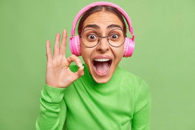 Close-up foto de feliz surpresa jovem fazendo gesto de ok mantém a boca bem aberta ouve transmissão de áudio em fones de ouvido usa óculos e gola olímpica isolada sobre a parede verde