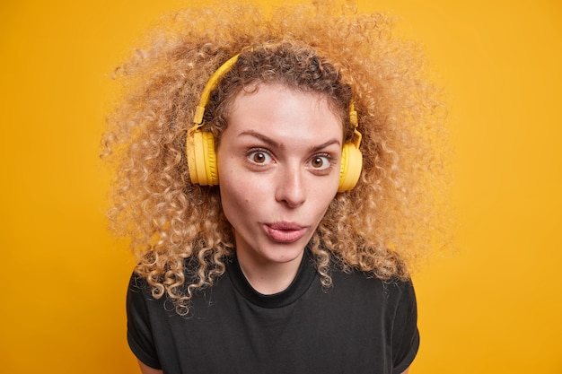 Close-up foto de uma bela adolescente cacheada usando fones de ouvido sem fio nas orelhas aprecia a qualidade do som ouve música mantém os lábios dobrados vestida com uma camiseta preta casual isolada sobre a parede amarela