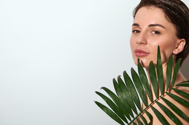 Close-up horizontal de uma linda mulher nua linda atraente com folha de palmeira exótica, posando contra um fundo branco com espaço de cópia. conceito de uso de cosméticos orgânicos naturais para cuidados com a pele