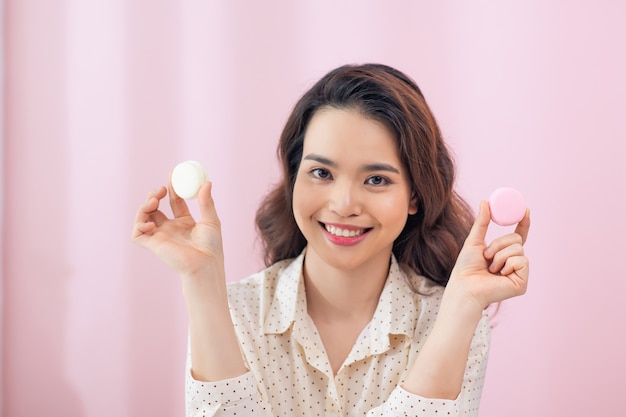 Close-up jovem mulher asiática comer macarons de bolo contra a parede rosa.