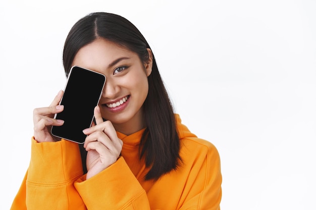 Close-up linda garota asiática com capuz laranja esconder o rosto atrás do smartphone, mostrando a tela do celular, promover o aplicativo, sorrindo alegre