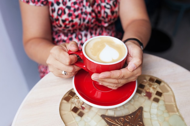 Close-up lindas mãos femininas abraçam o copo vermelho com bebida quente. Mãos de mulher segurando uma xícara de café em um café