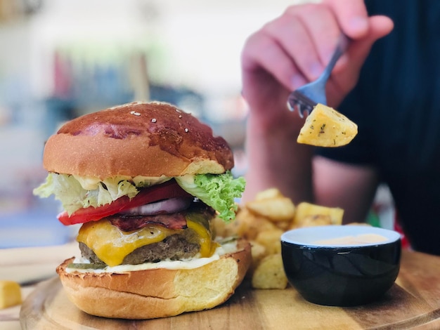 Foto close-up de la mano sosteniendo la patata en el tenedor en la mesa al lado de una gran hamburguesa