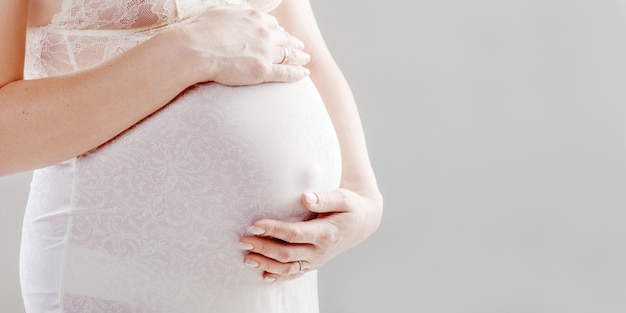 Close-up na barriga de grávida. Mulher esperando um bebê com uma barriga segurando as meias do bebê. Botinhas de bebé recém-nascido nas mãos de mães. Cor branca.