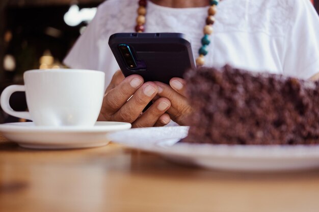 Close-up na mão de uma mulher sênior usando telefone celular enquanto relaxa para uma pausa no café com bolo de chocolate e xícara de café.