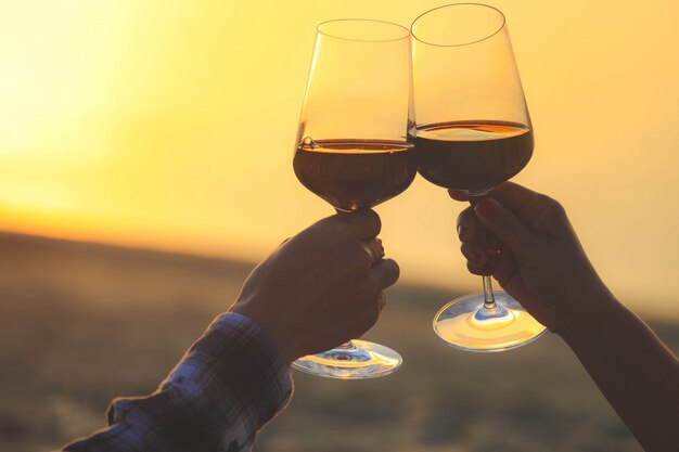 Close-up nas mãos segurando copos de vinho tinto na praia durante o pôr do sol, conceito de celebração