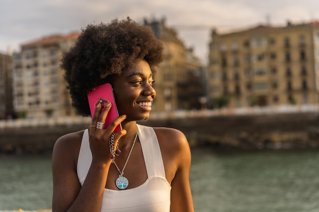 Close-up-Porträt einer abgelenkten und lächelnden jungen afrikanischen Frau, die während des Sonnenuntergangs am Meer mit dem Telefon spricht