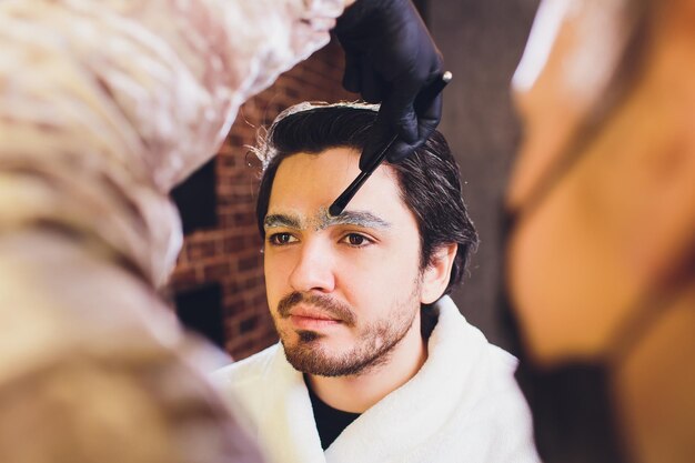 Close-up del proceso de procedimiento de enhechamiento en la barbería Barbero profesional corrigiendo la forma de las cejas con hilos a un joven sonriente sentado en una silla Concepto de cuidado de cejas