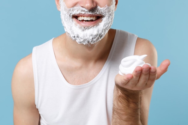 Close-up recortado de homem barbudo no rosto de camisa branca coberto com espuma de barbear isolada no retrato de estúdio de fundo pastel azul. Conceito de procedimentos cosméticos de saúde de cuidados com a pele. Simule o espaço da cópia.