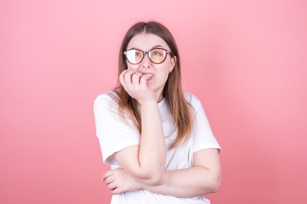 Close Up retrato de una adolescente preocupada en camiseta blanca mordiéndose las uñas aislado sobre fondo de color rosa