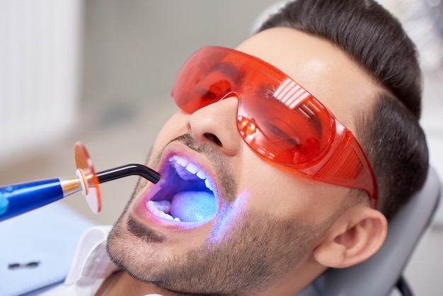 Close up retrato de un apuesto joven con gafas protectoras obteniendo el relleno de los dientes en la clínica dental profesionalismo seguridad salud medicina sonrisa tratamiento odontología.