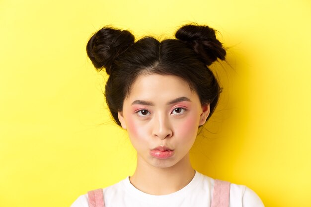 Close Up retrato de chica asiática de mal humor haciendo pucheros y mirando molesto a la cámara, de pie con maquillaje glamour y moños en amarillo
