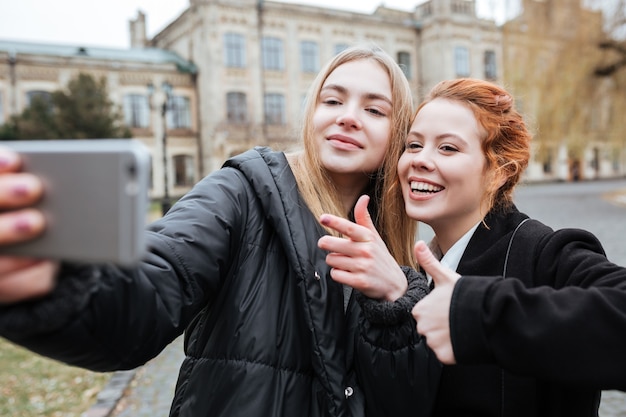Close Up retrato de dos adolescentes sonrientes tomando selfie y mostrando los pulgares para arriba gesto al aire libre