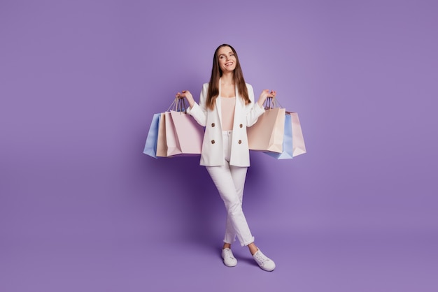 Close Up retrato de encantadora dama coqueta mantenga muchas bolsas de compras con las piernas cruzadas posando en la pared violeta