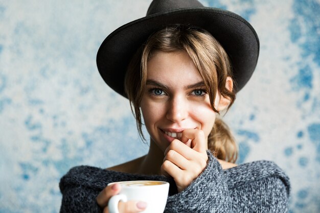 Close Up retrato de una hermosa joven vestida con suéter y sombrero sobre la pared azul, sosteniendo una taza de café caliente