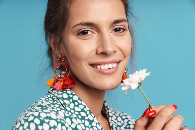 Close Up retrato de una hermosa joven vistiendo camisa y aretes brillantes que se encuentran aisladas sobre la pared azul, sosteniendo manzanilla