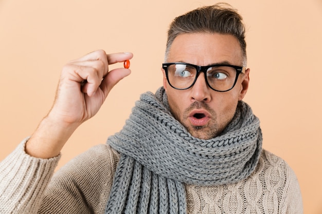 Close Up retrato de un hombre sorprendido vestido con suéter y bufanda que se encuentran aisladas sobre la pared beige, mostrando la cápsula de la píldora