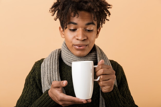 Close Up retrato de un joven africano feliz vestido con ropa de otoño aislado sobre pared beige, sosteniendo una taza