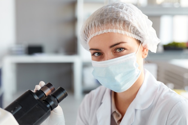 Close Up retrato de joven científica con mascarilla y mirando a cámara mientras trabaja con microscopio en laboratorio médico, espacio de copia arriba