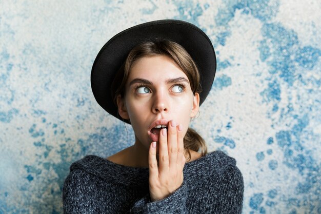 Close Up retrato de una joven sorprendida vestida con suéter y sombrero sobre la pared azul, mirando a otro lado