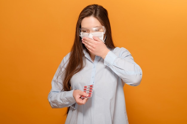 Close-up retrato de mujer joven en amarillo anaranjado en máscara médica protectora con síntomas de coronavirus, pastillas en las manos, tos y fiebre, pandemia de coronavirus