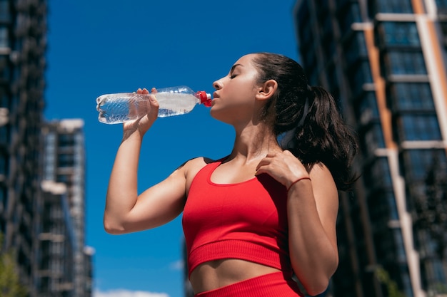 Close-up retrato de mujer joven con cabello castaño vistiendo ropa deportiva roja tomando descanso y bebiendo agua de plástico. Su mano izquierda con brazalete rojo tocando su pecho.