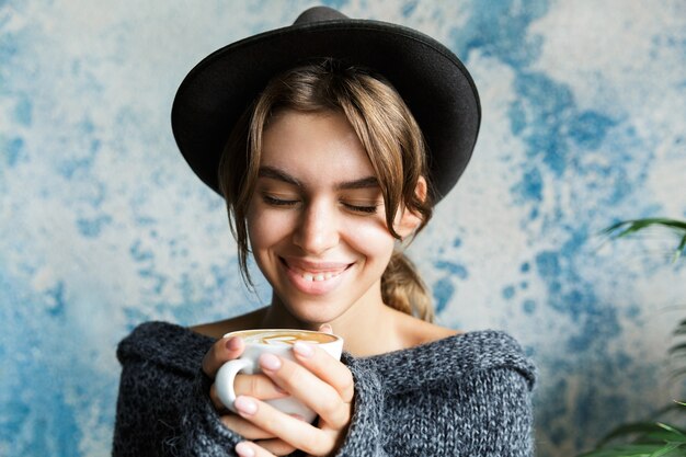 Close Up retrato de una mujer joven sonriente vestida con suéter y sombrero sobre la pared azul, sosteniendo una taza de café caliente