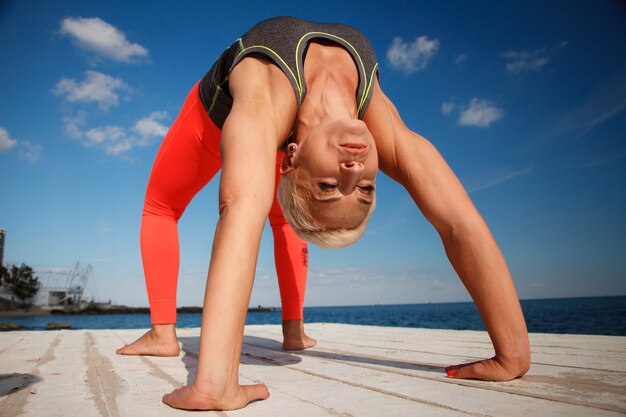 Close-up retrato de mujer rubia adulta con corte de pelo corto practica yoga en el muelle contra el fondo del mar y el cielo azul