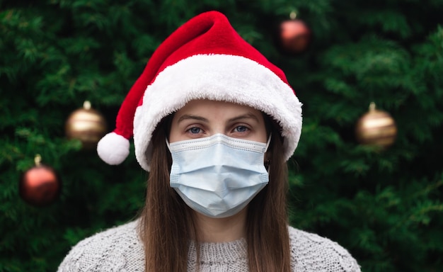 Close up Retrato de mujer con un sombrero de santa claus y una máscara médica con emoción. En el contexto de un árbol de Navidad. Pandemia de coronavirus