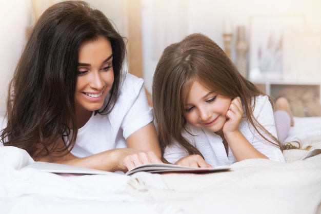 Close-up retrato de sonriente madre e hija leyendo en la cama temprano en la mañana en el blanco escandinavo