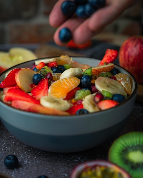 Foto close-up se a mão adicionando frutas à salada de frutas em uma tigela na mesa