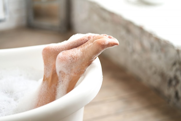 Close-up tiernos pies femeninos en una bañera blanca con espuma de jabón. Mujer disfrutando de un baño en un baño luminoso