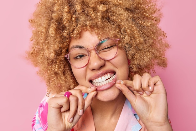 Close-up tiro de cabelo encaracolado jovem usa fio dental para limpar os dentes de comida pisca o olho usa óculos transparentes poses contra fundo rosa. higiene oral e conceito de cuidados com os dentes