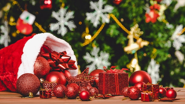 Close-up tiro de caixas de presente presentes de esfera estrela itens decorativos vermelhos derramando chapéu de Natal na mesa de madeira na frente da bela totalmente decoração com flocos de gelo meias pinheiro no fundo desfocado.