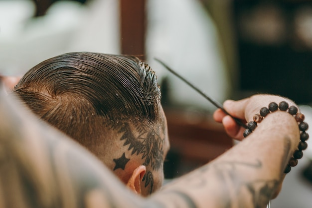 Close-up tiro de homem cortando o cabelo da moda na barbearia. O cabeleireiro masculino em tatuagens atendendo o cliente.